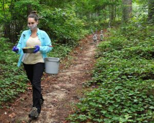 A masked person walks along a forest path with a bucket of lemur food, lemurs follow several feet behind