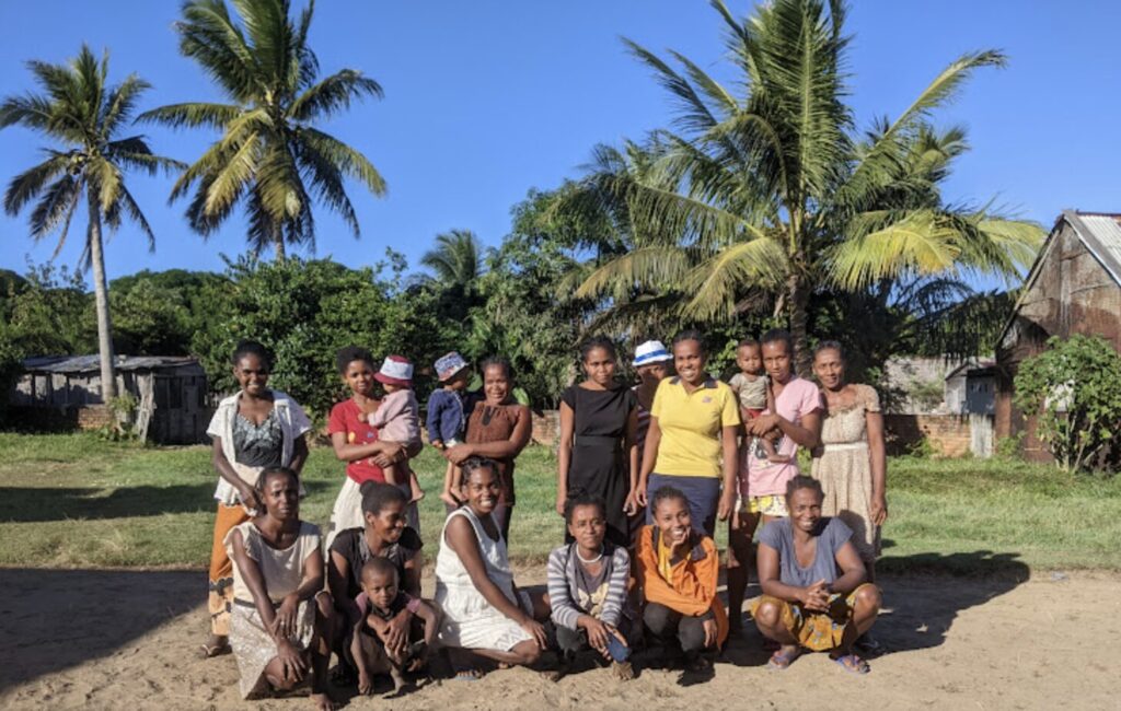 A group of women, some holding children, stand in front of palm trees and a bright blue sky in Madagascar.