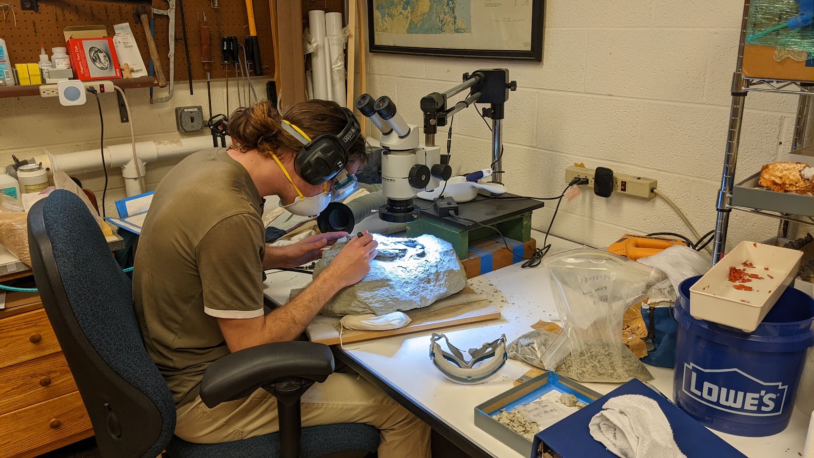 A Duke student with dark brown hair tied in a ponytail, ear protection, mask, and safety glasses bends over a workbench gently chisels at a rock to expose the fossil within.