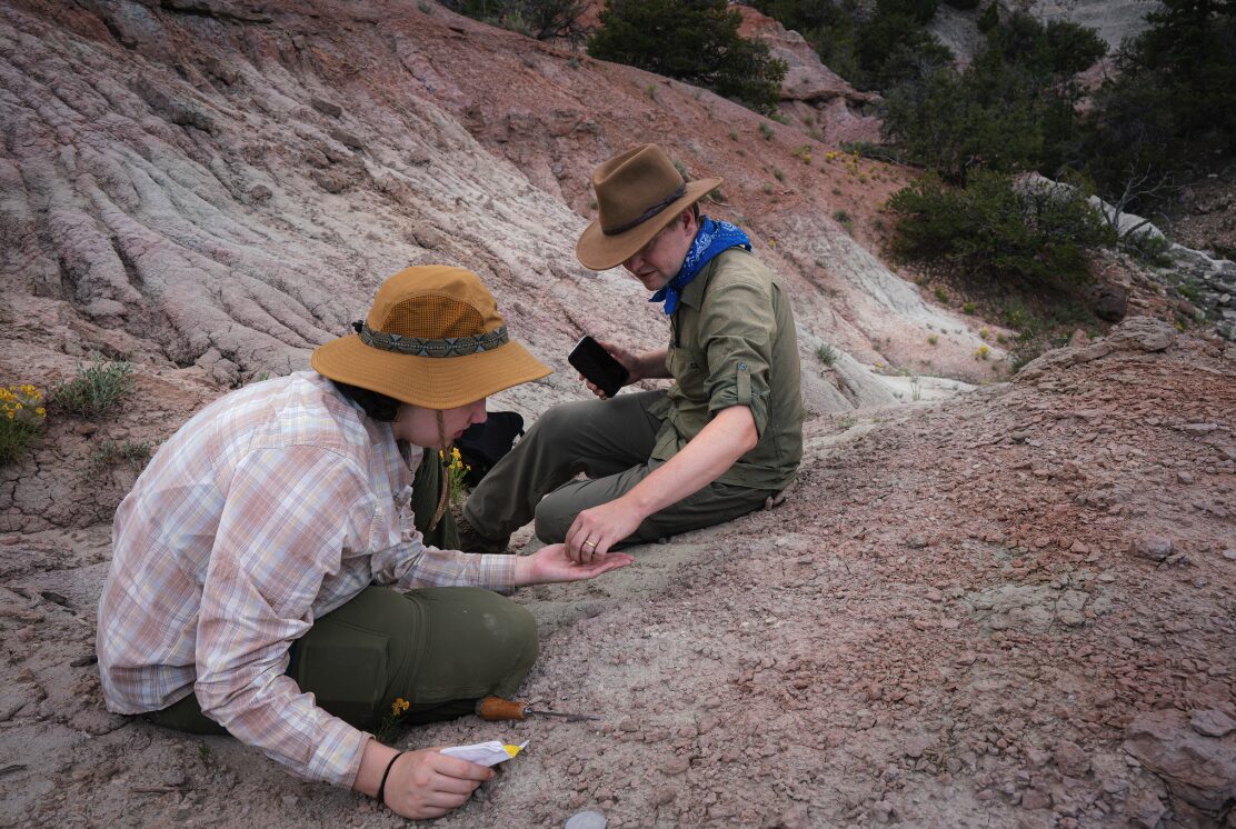 Two paleontologists wearing field hats examine a tiny fossil in rolling rocky cliffs shaded red, pink, and gray.