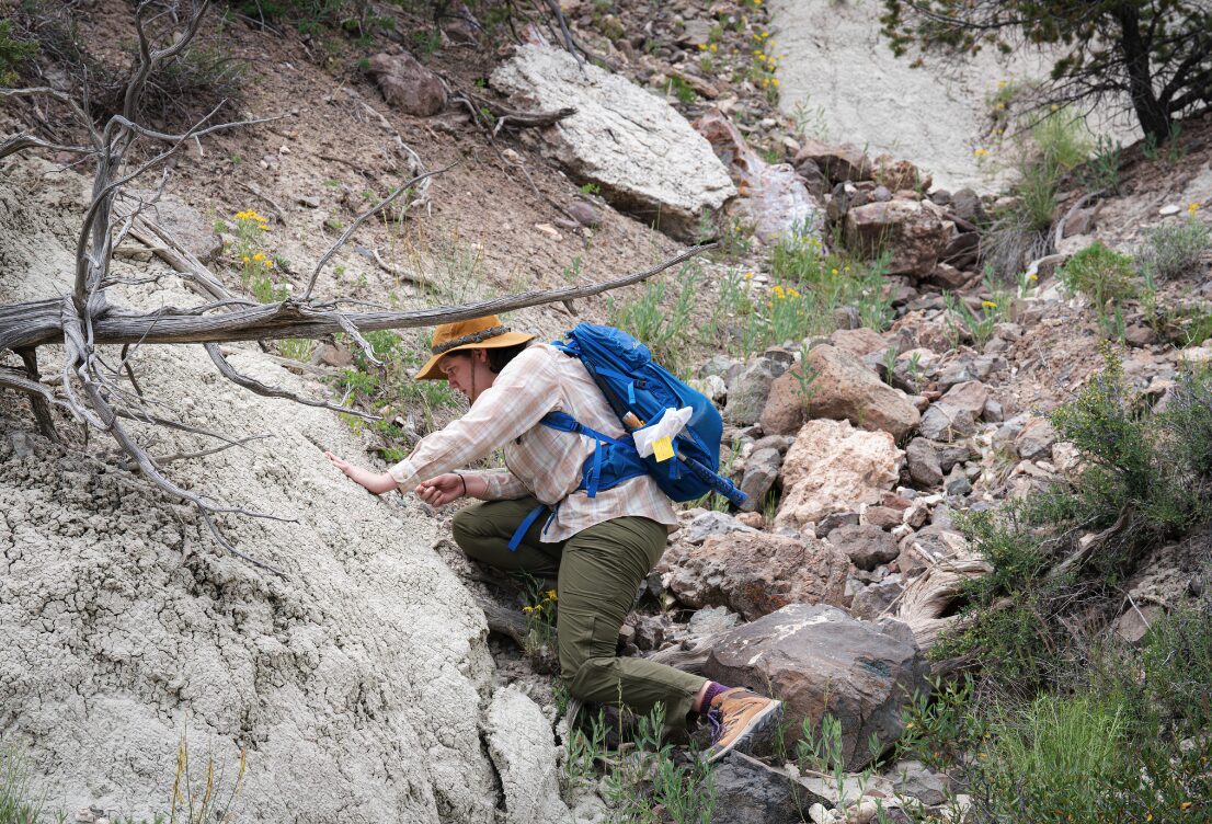 An undergraduate paleontologist wearing a field hat, field pants, backpack, and checked shirt chisels into a rock face looking for fossils.