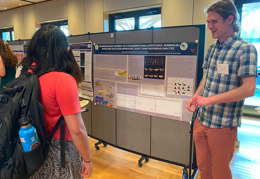 An undergraduate wearing a blue checked shirt, orange slacks, and name tag stands next to his research poster talking with a fellow student. The research poster is titled "Examining Dietary Diversity in a Paleogene Hyrax from the Fayum Depression, Egypt." 