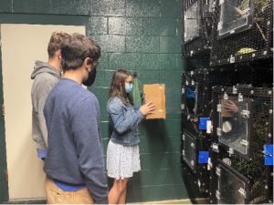 Three students holding cardboard structure in front of mouse lemur cages