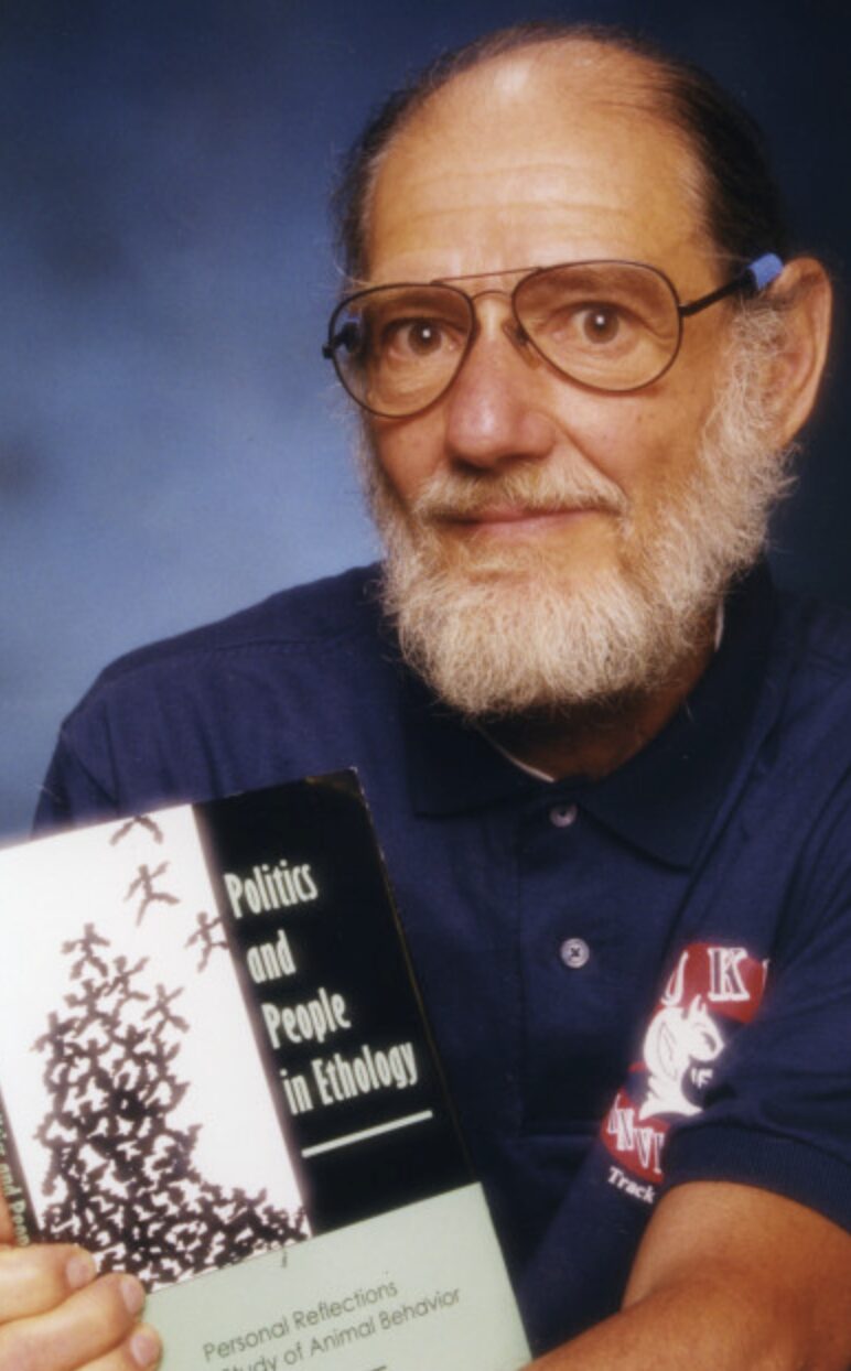 Peter Klopfer, wearing glasses and a black short-sleeved shirt with "Duke" on the left chest, holds his memoir titled "Politics and People in Ethology."