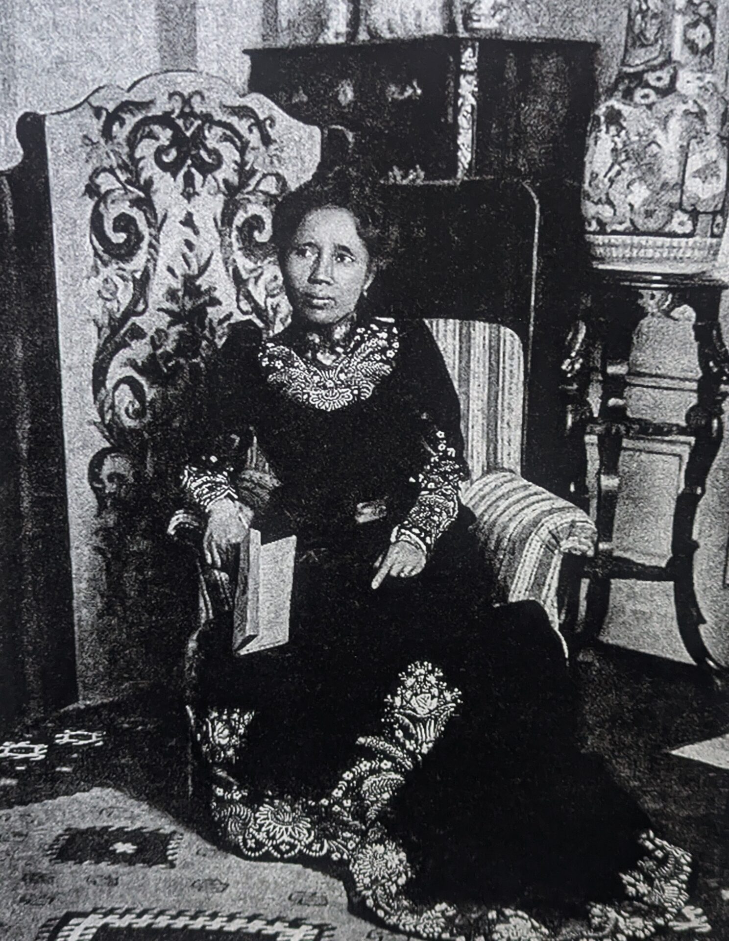 A grainy black and white photo of a Malagasy queen sitting in a striped chair against an ornate background.